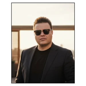 Homem com óculos escuros e terno preto ao pôr do sol.